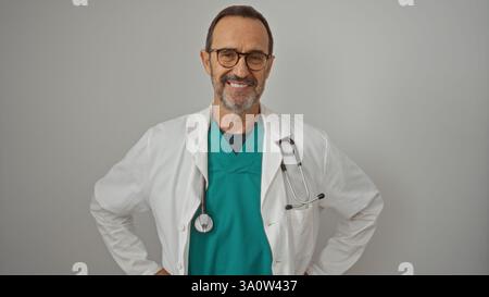 Reifer hispanischer Arzt lächelt selbstbewusst in grünen Peelings mit Stethoskop vor einem weißen Wandhintergrund und verkörpert Wärme und Professionalität Stockfoto