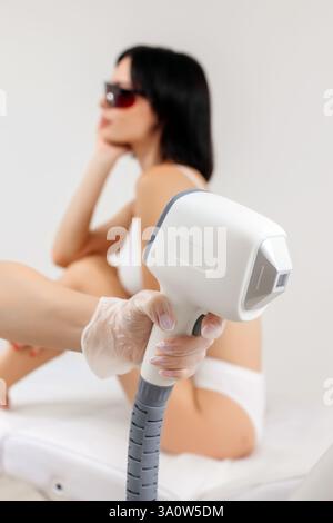 Körperpflegekonzept. Laserhaarentfernung. Junge weiße Frau in Schutzbrille liegt auf dem Sofa, weibliche Kosmetikerin, die Laser-Haarentfernung macht Stockfoto