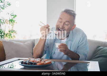 Glücklicher reifer Mann, der tagsüber Sushi und Sojasauce zu Hause in lässiger Kleidung genießt und Freizeitaktivitäten vorstellt Stockfoto