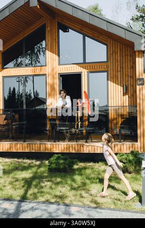 Mutter und Tochter genießen schöne Zeit in einer modernen Hotelhütte. Frau spielt mit Mädchen und rotem Spielzeugflugzeug in der Nähe eines Holzhauses. Urlaub und Familie Stockfoto