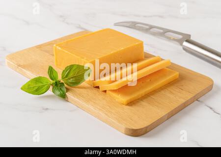 Sheddar-Käsescheiben auf einem Schneidebrett. Ganzer Käse auf einem hölzernen Schneidebrett und Messer über der Marmorarbeitsfläche. Milchproduktkonzept. Stockfoto