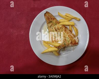 Ein Teller mit gegrilltem Kabeljaufilet mit goldenen Pommes frites, mit Olivenöl getränkt und mit Kräutern garniert, serviert in einer spanischen Mahlzeit. Stockfoto