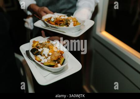 Ein Kellner serviert in einem schwach beleuchteten Restaurant zwei wunderbar gedeckte Gerichte mit lebhaftem Gemüse und Belägen. Stockfoto