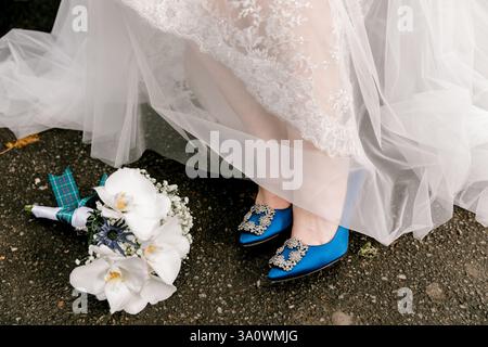 Die wunderschöne Braut steht auf strukturiertem Boden, trägt auffällige blaue Schuhe und hält einen zarten weißen Blumenstrauß. Stockfoto