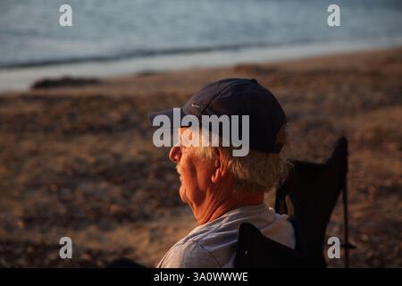 Mann sitzt am Strand von Vouliagmeni und beobachtet den Sonnenuntergang am Meer Athen Attika Griechenland Stockfoto