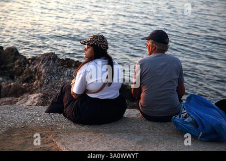 Vater und Tochter sitzen auf einem Felsen und beobachten den Sonnenuntergang am Vouliagmeni Beach Athen Attika Griechenland Stockfoto