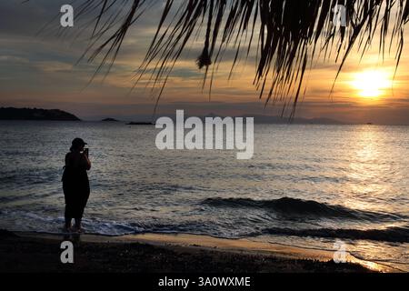 Frau, die im Meer steht und den Sonnenuntergang beobachtet, fotografiert mit Handy Vouliagmeni Beach Attica Griechenland Stockfoto