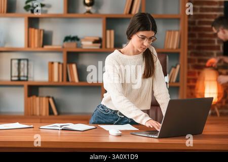 Eine junge Frau in Brille arbeitet drinnen. Stehtisch mit Laptop. Stockfoto