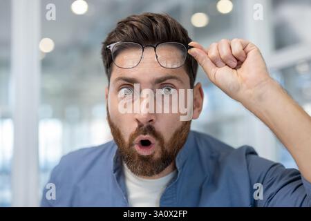 Nahaufnahme eines Mannes mit Bart und blauem Hemd, der überrascht aussieht und seine Brille auf einem unscharfen Hintergrund hebt Stockfoto