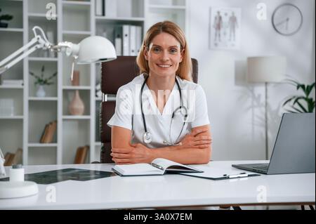 Am Tisch sitzen. Die junge Ärztin im weißen Mantel ist drinnen. Stockfoto