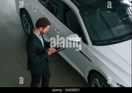 Draufsicht, mit Tablet. Mann in schwarzem Anzug ist drinnen im Autohändler in der Nähe des Autos. Stockfoto