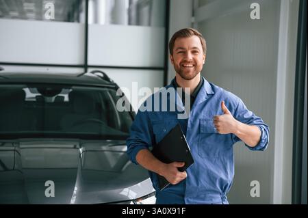 Lächelnd, stehend mit Notizblock in der Hand. Ein Mann in blauer Uniform arbeitet im Autodienst. Stockfoto