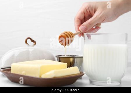 Frau mit Honiglöffel, Nahaufnahme. Frische Milch im Glas und Butter auf weißem Fliesentisch Stockfoto