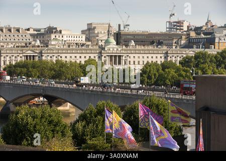 Fußgänger auf der Waterloo Bridge, London, sehen die historischen Gebäude und Bauten entlang der Themse. Flags sind sichtbar. Stockfoto