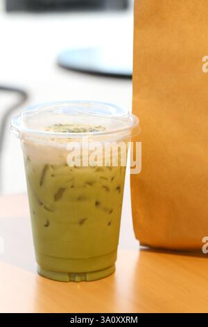 Eine Nahaufnahme eines erfrischenden, eiskalten Matcha-Latte in einer klaren Tasse mit Strohhalm, der neben einer braunen Papiertüte auf einem Holztisch in einer hellen Café-Atmosphäre sitzt Stockfoto