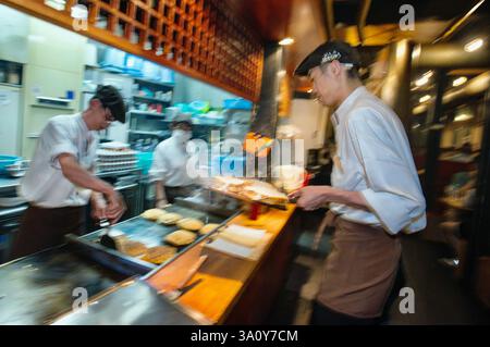 Yoshi Miyahara (R) führt Okonomiyaki-Pfannkuchen, die am 20. Juli vom Küchenchef Masanobu Nishikura (L) im Hauptgeschäft von Boteju in Osaka, Japan, auf der Grillplatte zubereitet werden Stockfoto