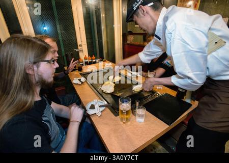 Yoshi Miyahara (R) liefert Okonomiyaki-Pfannkuchen am 20. Juli 2017 im Hauptgeschäft von Boteju in Osaka, Japan, auf eine Grillplatte für Kunden aus Dänemark. Stockfoto