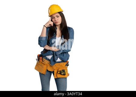 Eine junge Frau mit gelbem Schutzhelm und Denim-Hemd, mit einem Werkzeuggürtel und einem nachdenklichen Ausdruck. Sie reibt sich die Stirn, als ob sie es wäre Stockfoto