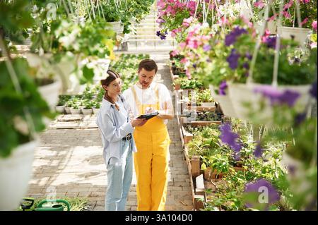 Orangefarbene Uniform. Floristen-Mann und -Frau arbeiten in einem hellen Gewächshaus zusammen. Stockfoto