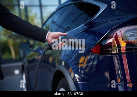 Hand hält das Ladegerät, Nahansicht. Der Geschäftsmann steht draußen in der Nähe seines Elektroautos. Stockfoto