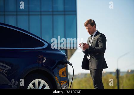Geschäftsgebäude dahinter. Ein Mann im Anzug steht draußen neben seinem Elektroauto. Stockfoto