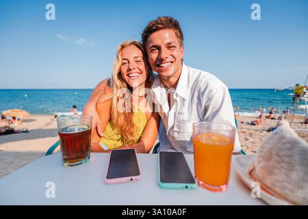 Fröhliches Paar genießt erfrischende Getränke auf einer sonnigen Strandterrasse mit atemberaubendem Meerblick, während es gemeinsam Spaß hat und schöne Erinnerungen während ihres herrlichen Sommerurlaubs schafft. Hochwertige Fotos Stockfoto