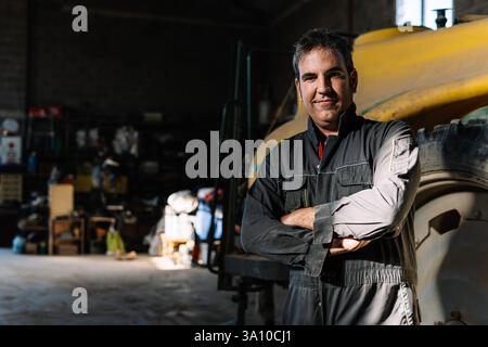 Ein selbstbewusster spanischer Bauer steht stolz in seiner Scheunenwerkstatt, die Arme gekreuzt, und sein Traktor steht hinter ihm, der den Geist der Landwirtschaft verkörpert Stockfoto