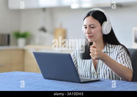 Asiatische Frau, die Laptop und Kopfhörer benutzt und Kaffee in der Küche zu Hause trinkt Stockfoto