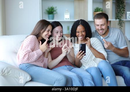 Hoffnungsvolle, glückliche, interrassische Freunde, die die Finger drücken und das Telefon auf einer Couch zu Hause checken Stockfoto