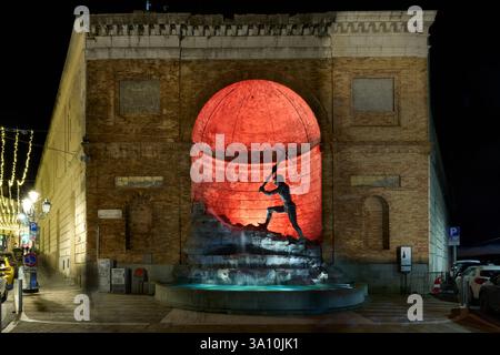 Nächtlicher Blick auf einen historischen Brunnen mit einer Statue, die von rotem Licht beleuchtet wird, vor einem alten Backsteingebäude in einer städtischen Umgebung. Stockfoto