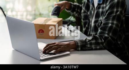 Nahaufnahme eines jungen Mannes, der einen Laptop benutzt, während er ein Paket für den Versand scannt. Stockfoto