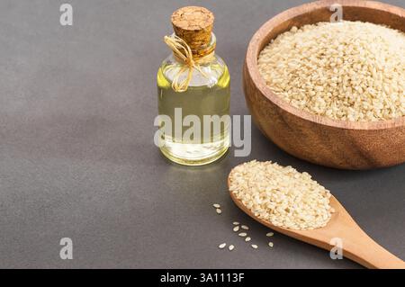 Glasflasche mit Sesamöl und rohen Sesamsamen in Holzlöffel und Schüssel auf rustikalem Tisch. Unbehacktes Sesamhintergrundkonzept mit Kopierraum Stockfoto