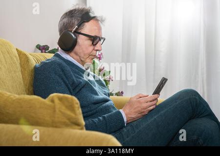 Senior-Mann mit schnurlosen Kopfhörern, Musik oder Podcasts, während er sein Smartphone verwendet. Moderner Lifestyle und digitale Konnektivität zu Hause. Stockfoto