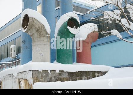 Drei große Lüftungsrohre - rot, grün und grau - stapeln sich vor einer blauen Industrieanlage mit Schnee und zeigen eine lebhafte urbane Szene in Kälte Stockfoto
