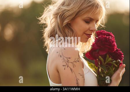 Seitenansicht. Die schöne junge Frau steht draußen und hält rote Blumen. Stockfoto