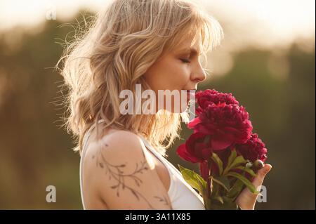 Seitenansicht. Die schöne junge Frau steht draußen und hält rote Blumen. Stockfoto
