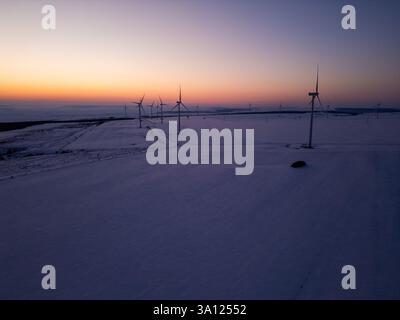 Luftaufnahme eines Windparks in einer schneebedeckten Landschaft bei Sonnenuntergang. Windturbinen stehen hoch gegen das warme Leuchten der untergehenden Sonne und erzeugen einen Stockfoto