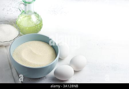 Backzutaten: Hellblaue Schüssel mit Grieß, frische weiße Eier, eine Glasschale mit Kristallzucker und eine Flasche Pflanzenöl auf einem weißen Stockfoto