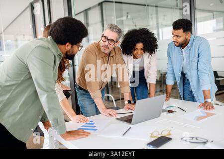 In einem modernen Büro versammelte sich ein Team von Fachleuten um einen Laptop, analysierte Daten und diskutierte Strategien. Eine vielfältige Gruppe, die an einem Geschäftsprojekt mit Dokumenten, Diagrammen und Technologie zusammenarbeitet Stockfoto