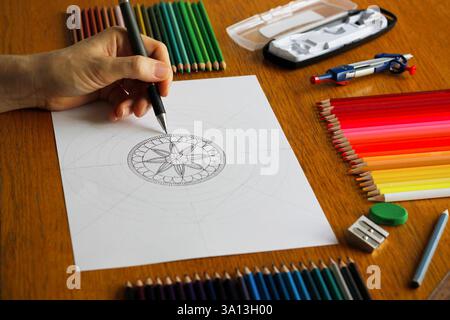 Linke Hand einer Kaukasierin, die ein Mandala-Muster auf weißem Papier auf einem Holztisch zeichnet, bunte Bleistifte und andere Zeichenutensilien in der Nähe. Stockfoto