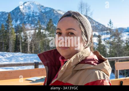 Eine 45-50-jährige Frau in warmen Winterkleidung, die auf einer Außenterrasse mit Blick auf schneebedeckte Berge und Wälder sitzt. Klares, sonniges Wetter schafft Stockfoto