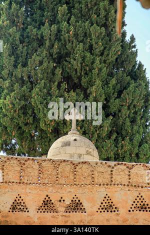 Wunderschönes Kreuz auf einer Steinkuppel, an einer Wand mit dekorativen Designs und hohen grünen Zypressen unter klarem Himmel, die Spiritualit darstellen Stockfoto