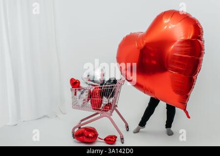 Großes rotes Herz mit rosa Wagen voller Ballons auf weißem Hintergrund. Fotozone mit Herzen für Liebhaber Stockfoto