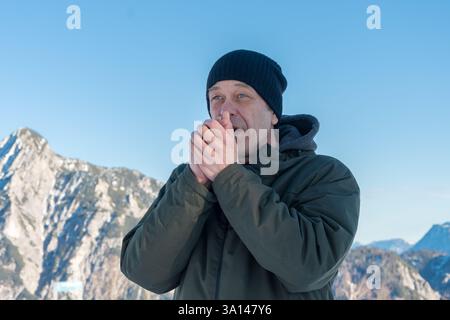 Ein älterer Mann, 50-55 Jahre alt in Winterkleidung, steht vor einem Hintergrund schneebedeckter Berge und blauem Himmel und wärmt seine Hände mit dem Atem Stockfoto