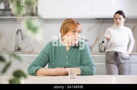 Ältere traurige Frau sitzt am Tisch in der Küche und hört Beleidigungen zu. Konzeptstrom des Missbrauchs Stockfoto