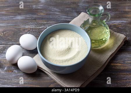 Eine blaue Keramikschale mit Grieß, drei frischen weißen Eiern und einer Glasflasche Pflanzenöl auf einem rustikalen Holztisch Stockfoto