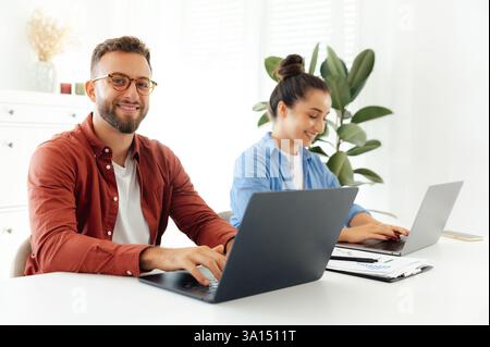 Ein Mann und eine Frau arbeiten zusammen an Laptops, lächeln und arbeiten zusammen. Zwei erfolgreiche multirassische Kollegen arbeiten an einem neuen Projekt in einem modernen Bürobereich in freundlicher Atmosphäre Stockfoto