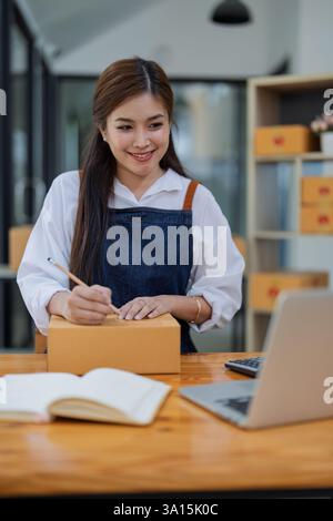 Junge Frau, die im Online-Geschäft tätig ist und mit Laptop-Computern arbeitet, Pakete für den Versand vorbereitet und Notizen der Kundenadresse an der Box macht. Stockfoto