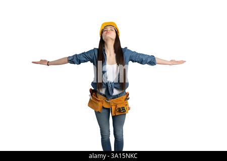 Junge Bauarbeiterin mit gelbem Schutzhelm und Werkzeuggürtel, stehend mit weit geöffneten Armen und geschlossenen Augen. Sie trägt einen Denim-Shir Stockfoto