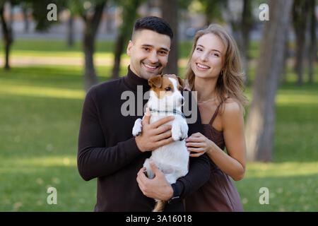 Ein Paar genießt einen sonnigen Tag in einem üppigen Park und hält seinen liebenswerten Hund. Ihr Lächeln strahlt Glück aus, während sie die Wärme der Natur genießen, cre Stockfoto